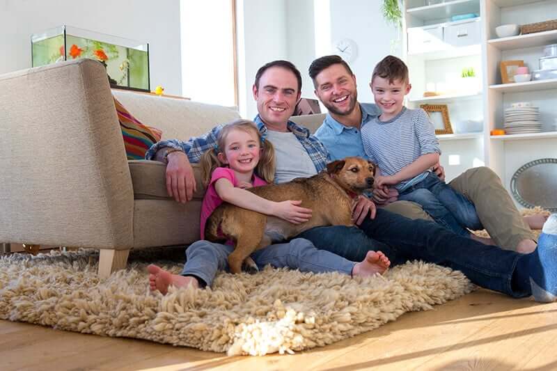Same sex male couple sitting on the floor in their living room with their son and daughter. Their pet dog is lying across them.
