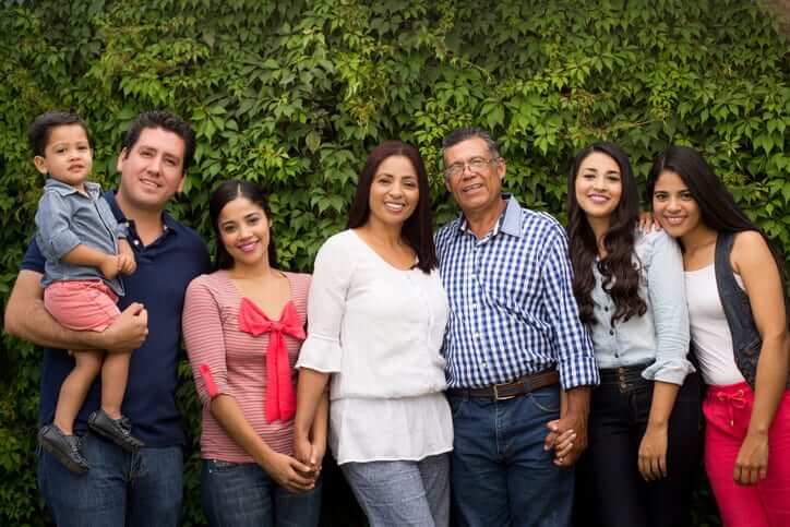 A multi-generation latin family of seven standing, holding hands and smiling at the camera in a horizontal medium shot outdoors.