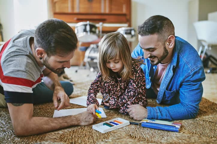 Gay parents assisting daughter with drawing at home. Homosexual couple and girl are spending leisure time in living room. They are wearing casuals.