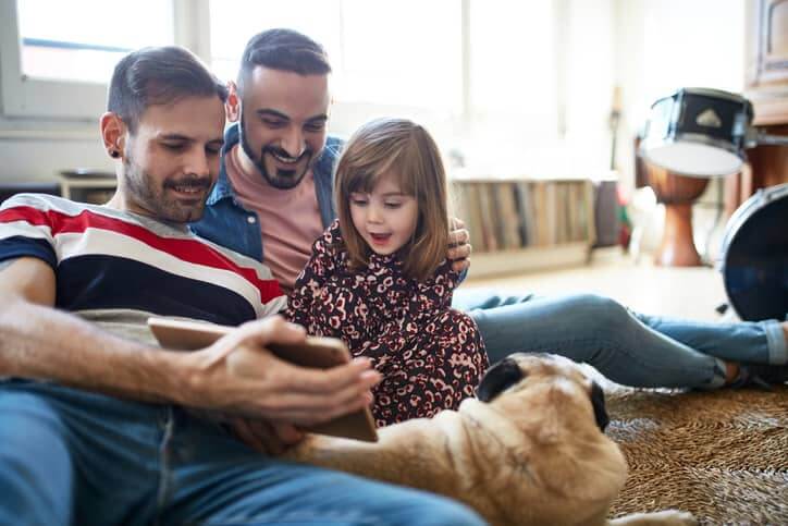 Smiling Spanish fathers enjoying weekend leisure with young daughter and dog as they use digital tablet together on living room floor.