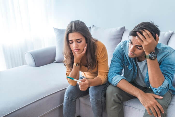 Sad stressed couple with pregnancy test sitting at home. Upset woman is looking in pregnancy test. Frustrated man is sitting next to her. Worried about the serious consequences