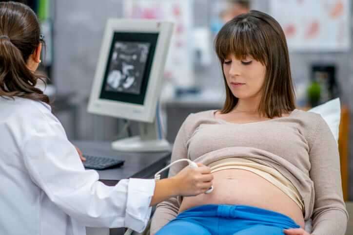 A female doctor and pregnant woman are inside a hospital room. The doctor is looking at a computer screen while performing an ultrasound.